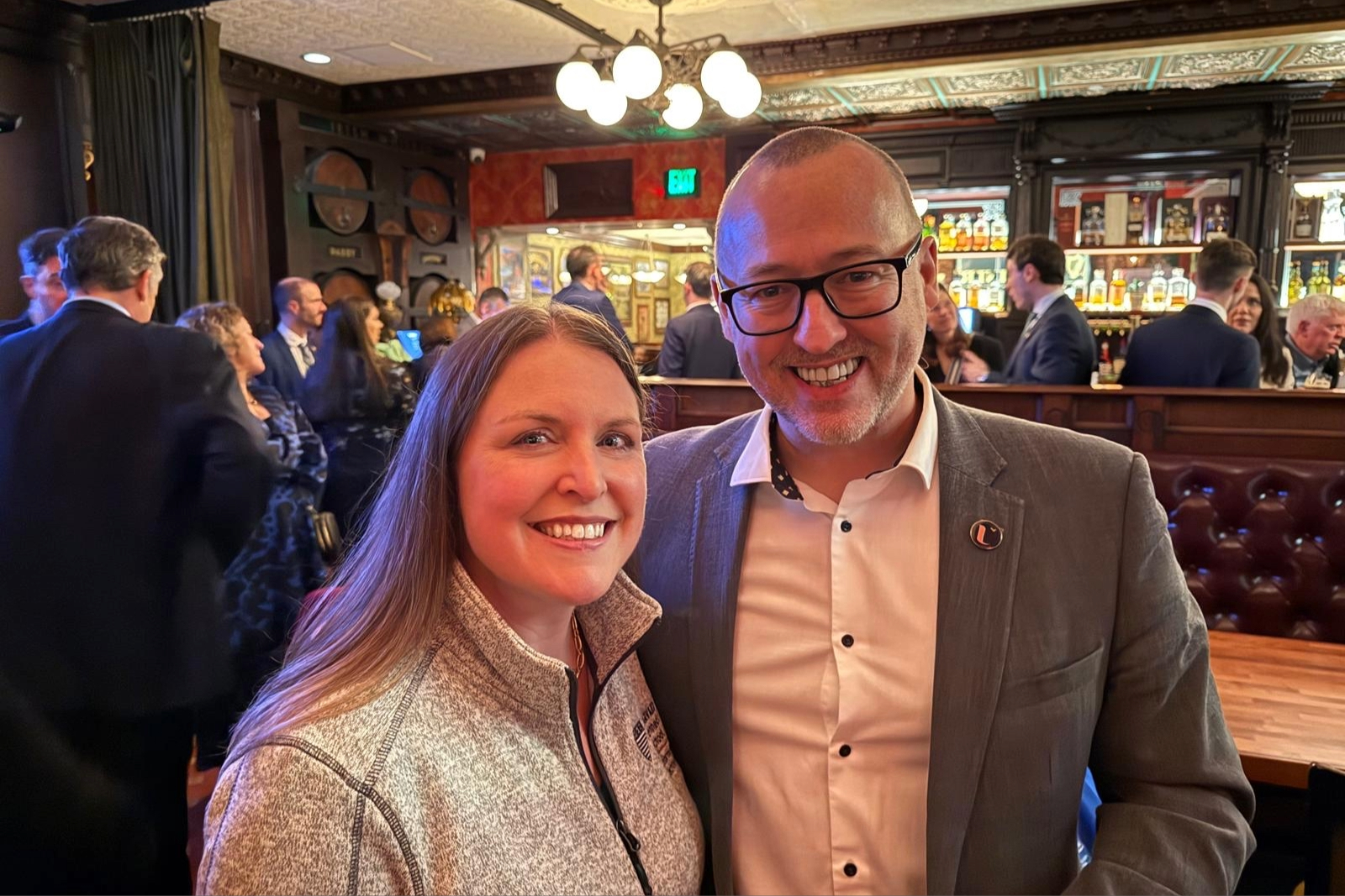 A man and woman smiling in a warmly lit bar with a lively crowd. People dressed in formal attire chat in the background near a stocked bar.